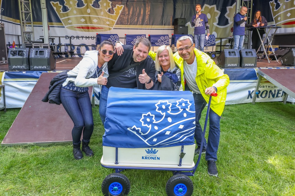 Vier Erwachsene lächeln und posieren mit einem dekorierten Wagen bei einem Outdoor-Event.