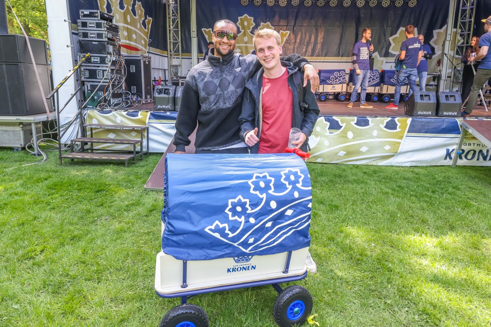 Zwei Männer stehen lächelnd neben einem kleinen Wagen bei einem Outdoor-Musikevent.
