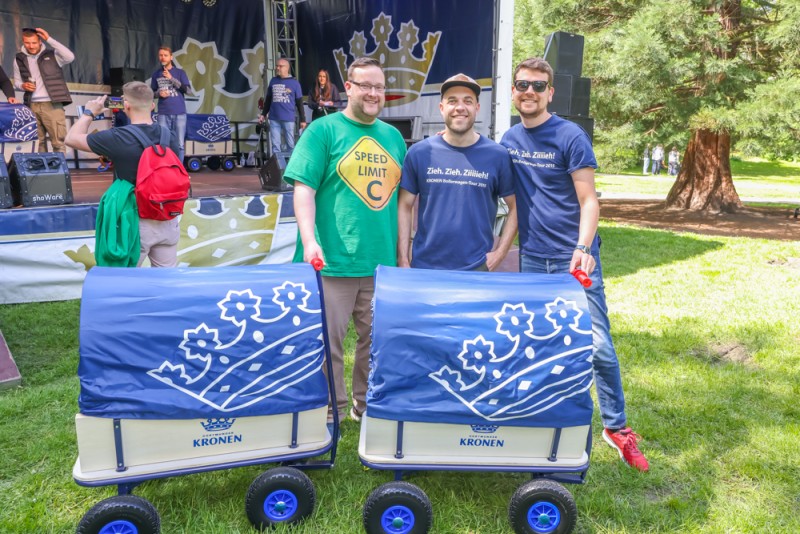 Drei Männer stehen draußen nebeneinander, zwei davon halten jeweils einen Bollerwagen.