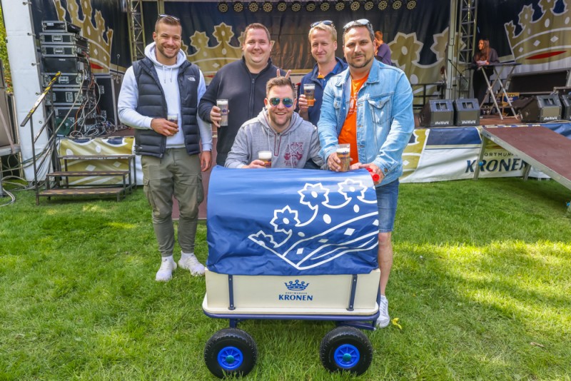 Fünf Männer stehen lächelnd mit Biergläsern neben einem Wagen auf einer Wiese.