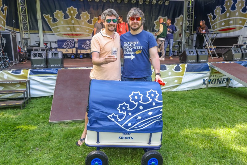 Zwei Männer stehen mit einem blau dekorierten Wagen vor einer Bühne im Freien.