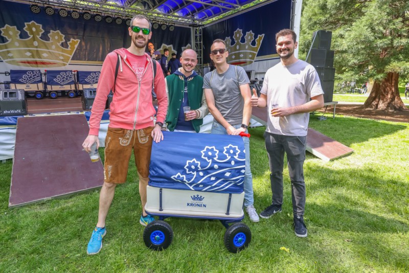 Vier Männer stehen lächelnd neben einem dekorierten Bollerwagen auf einer Wiese.