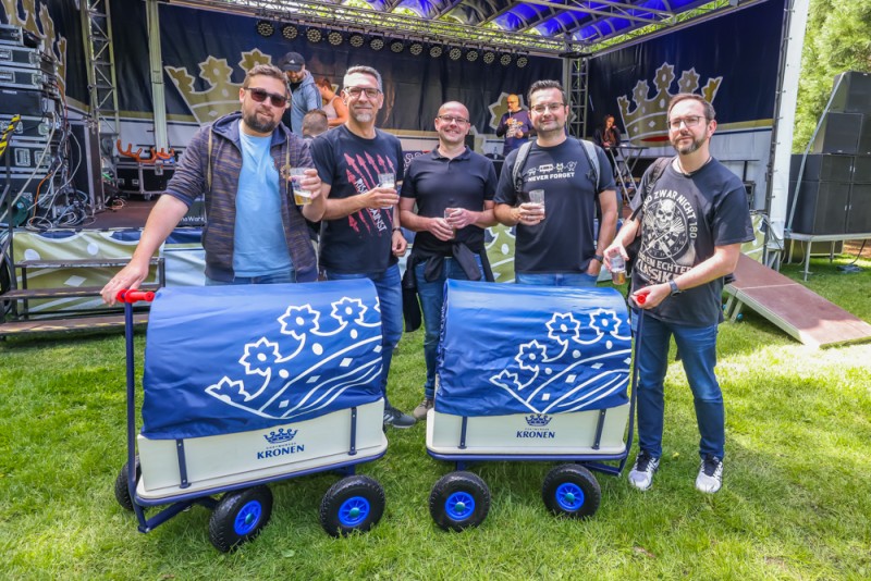 Fünf Männer stehen lächelnd vor einer Bühne und halten Getränke, zwei lehnen an Handwagen.