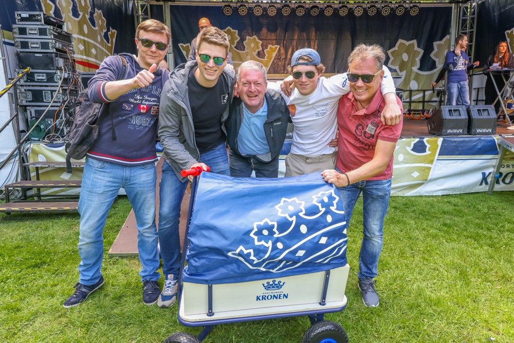 Fünf Männer lächeln und posieren auf einer Wiese mit einer Fahne vor einer Bühne.