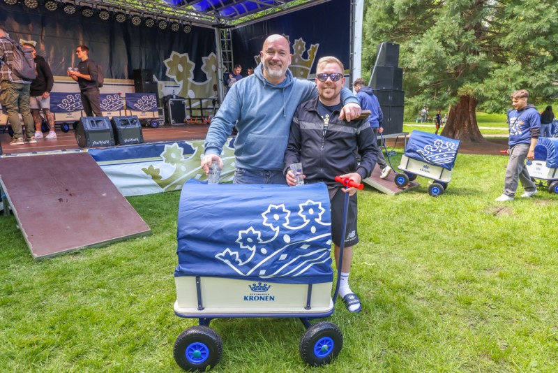 Zwei Männer stehen lächelnd auf einer Wiese, einer hält einen bemalten Wagen.