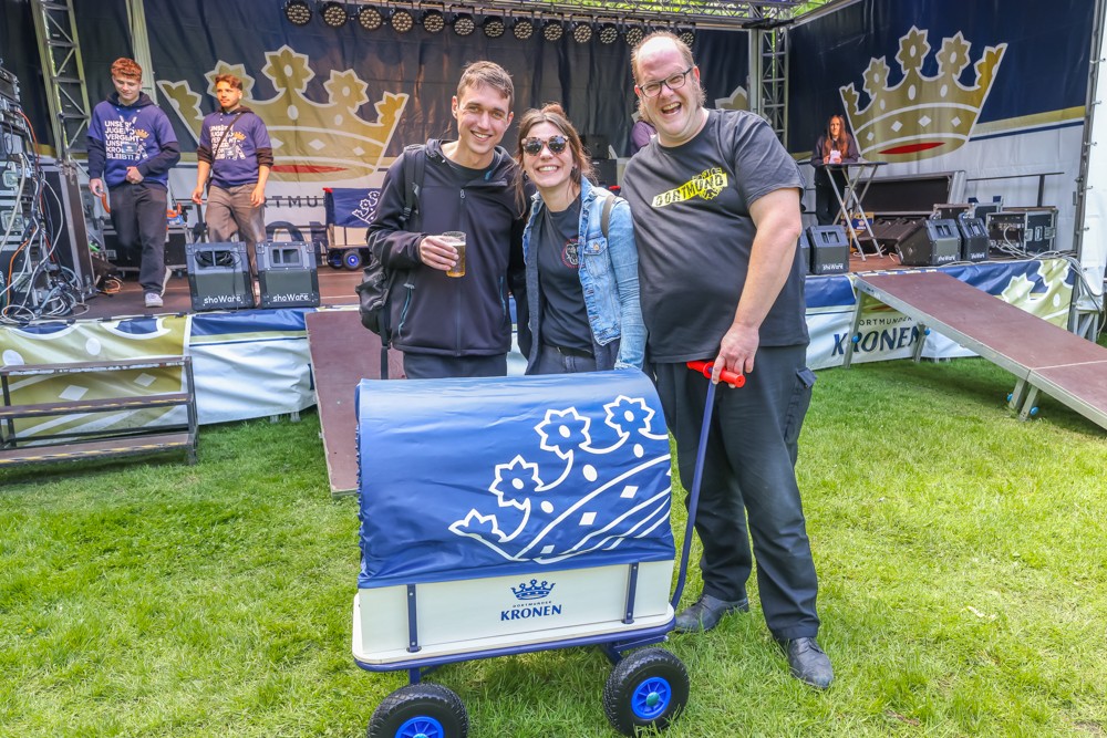 Drei Personen stehen lächelnd vor einer Bühne mit einem blauen Bollerwagen.