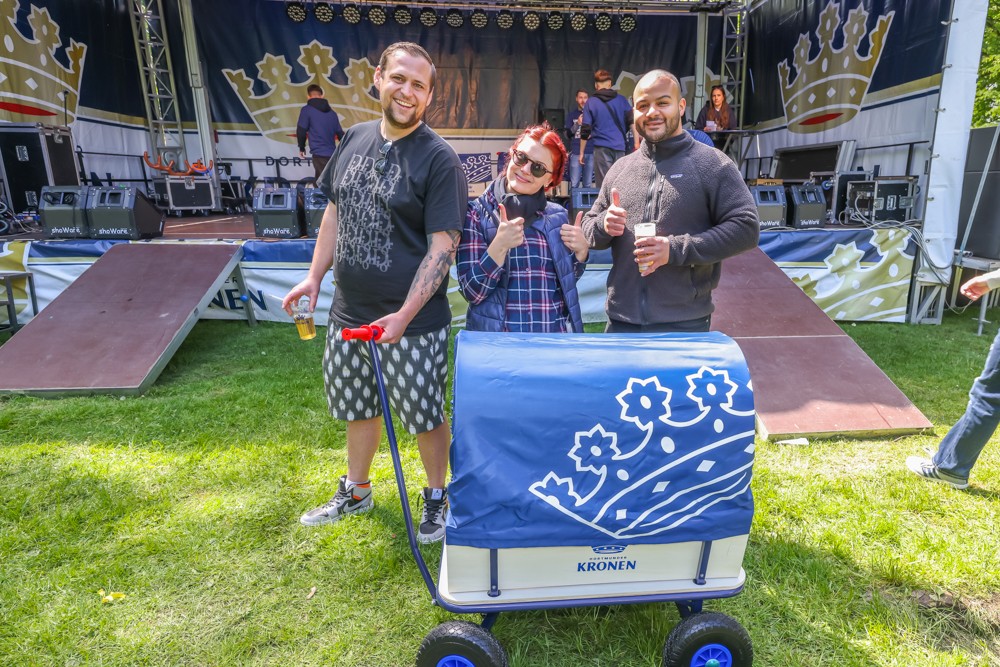 Drei Personen stehen lächelnd auf einer Wiese vor einer Festivalbühne, zwei zeigen Daumen hoch.