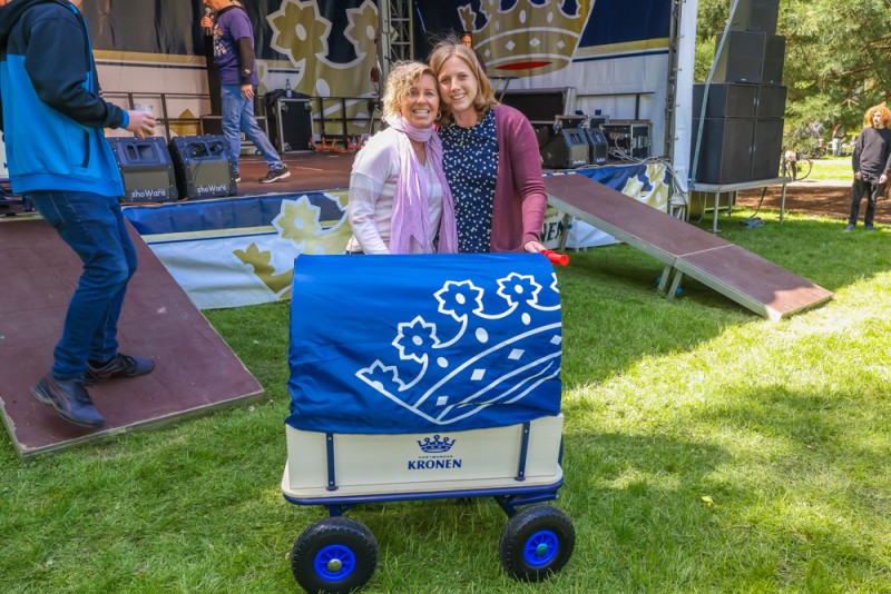 Zwei Frauen lächeln neben einem Handwagen bei einem Event im Freien.