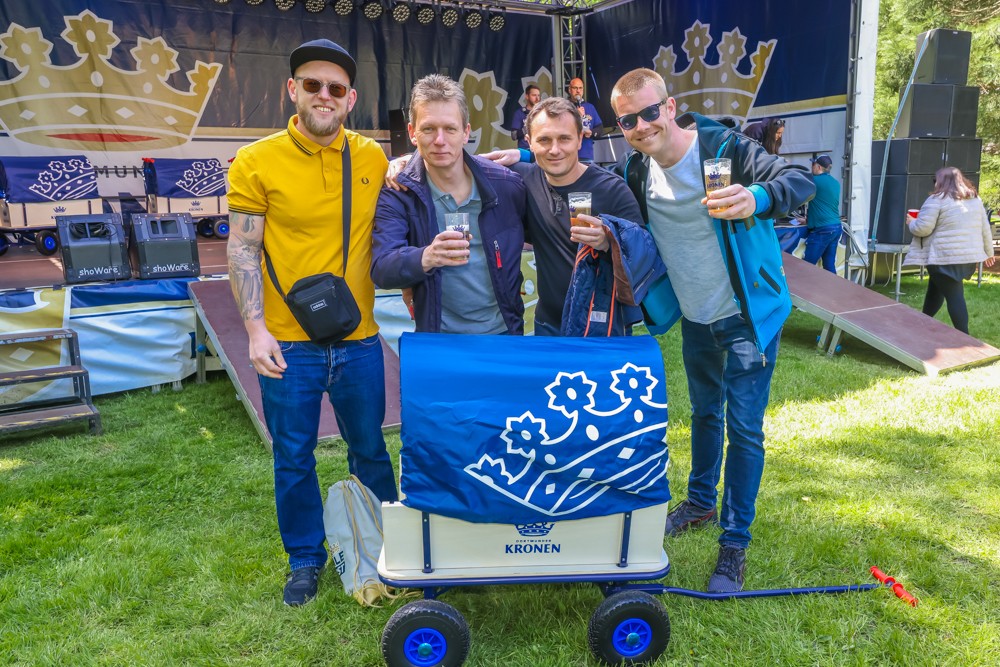 Vier Männer stehen lächelnd vor einer Bühne und halten Gläser. Einer zieht einen blauen Bollerwagen.