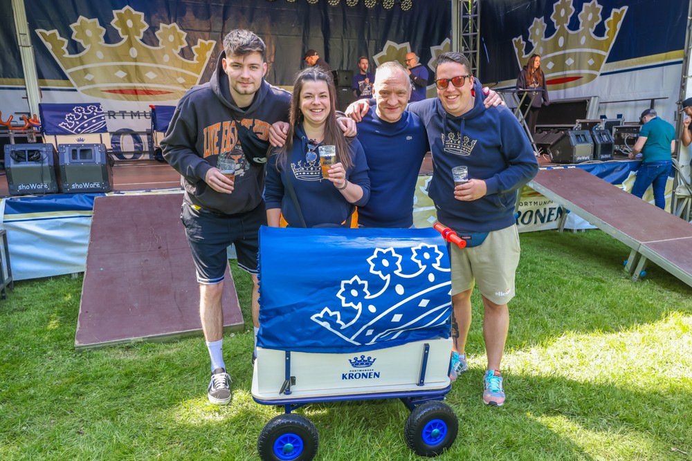 Vier Personen stehen lächelnd neben einem Bollerwagen auf einem Festival.