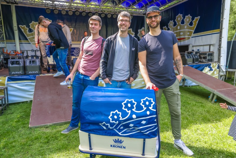 Drei Männer posieren lächelnd vor einer Bühne mit Musikinstrumenten im Freien. Sie halten ein blaues Banner mit grafischen Symbolen.