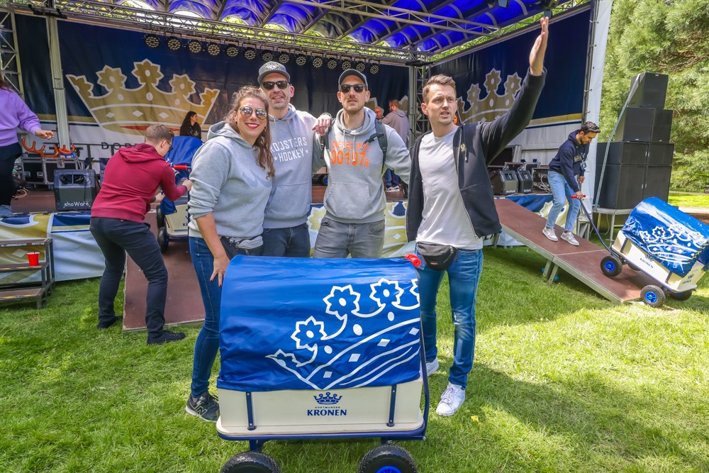 Vier Erwachsene stehen lächelnd auf einer Wiese vor einer Bühne neben einem blau dekorierten Wagen.