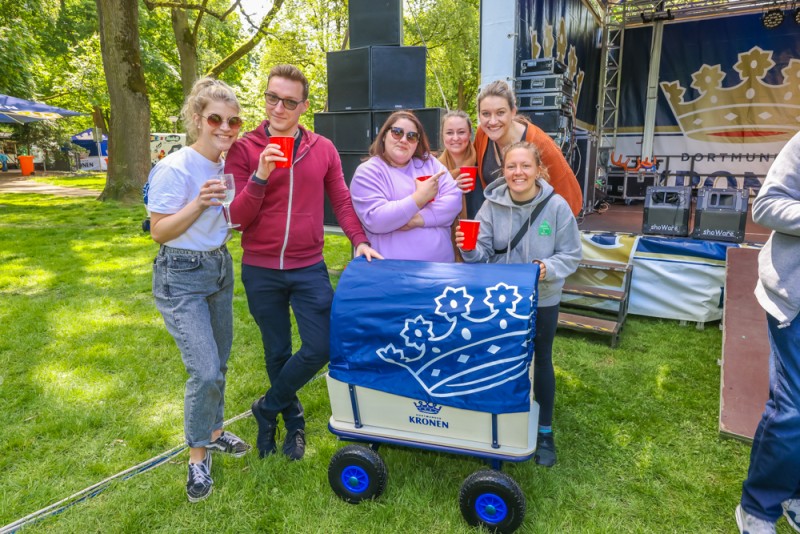 Sechs Personen stehen lächelnd im Park bei einem Event und halten rote Becher.