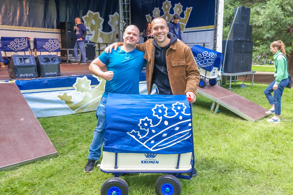 Zwei Männer stehen lächelnd neben einem blauen Bollerwagen auf einer Veranstaltung.