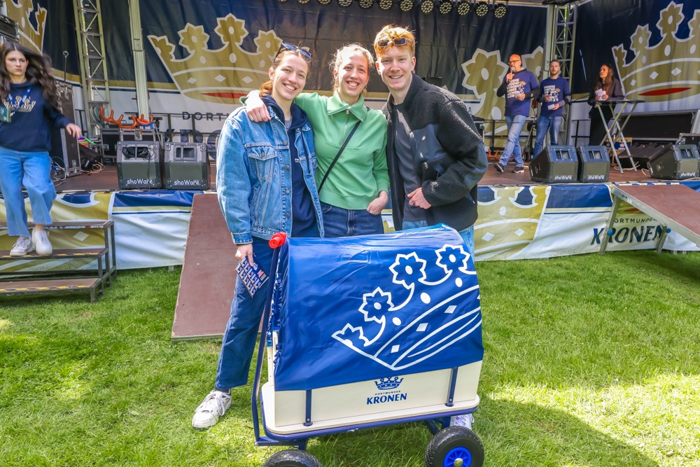 Drei Personen lächeln vor einer Festivalbühne, neben ihnen steht ein blauer Handwagen.
