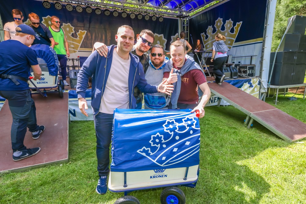 Vier Männer stehen lächelnd mit einem blauen Bollerwagen bei einem Festival.