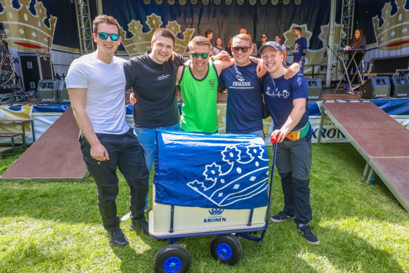 Fünf junge Männer lächeln neben einem blauen Wagen auf einer Veranstaltung.