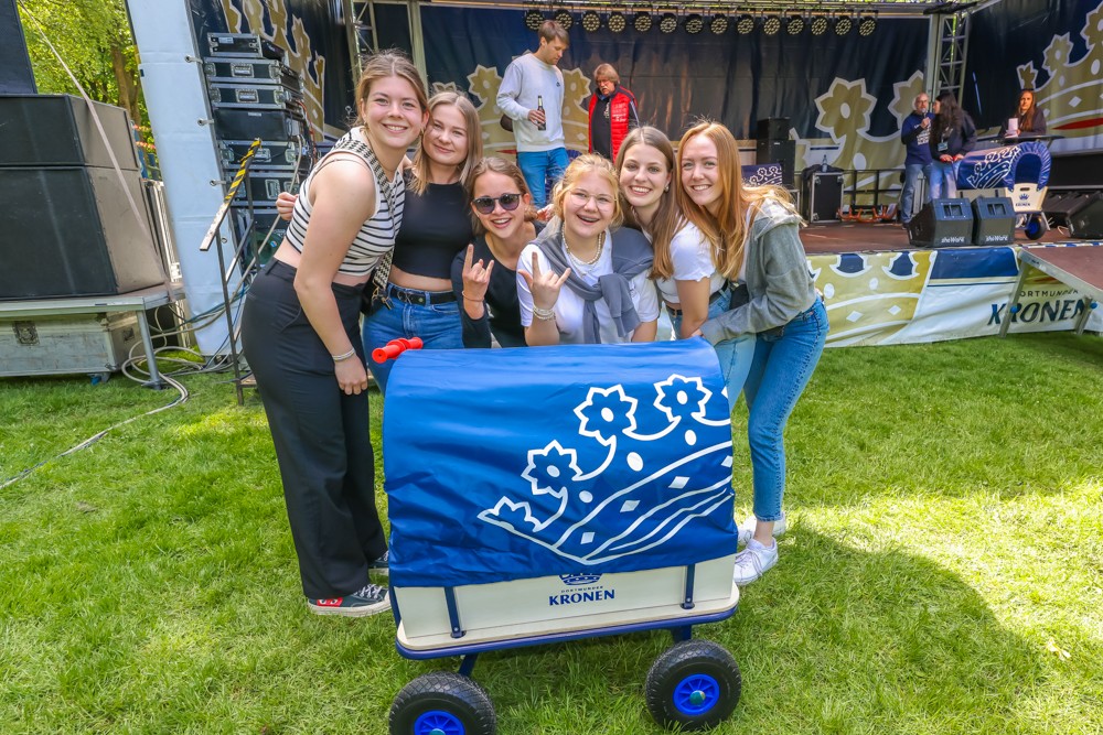 Sechs lächelnde Frauen stehen mit einem blauen Wagen vor einer Bühne im Park.