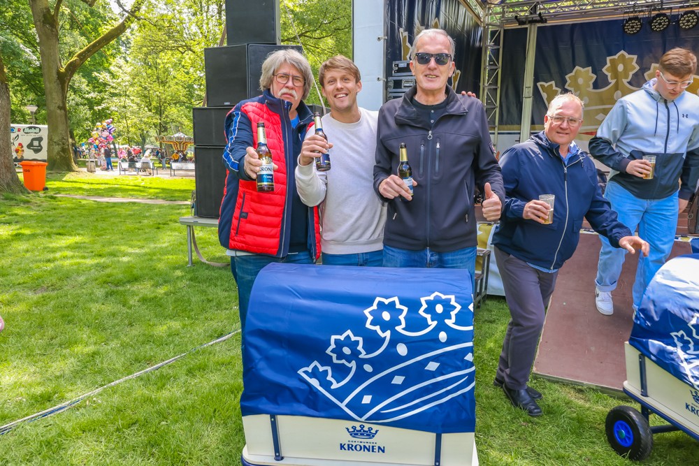 Vier Männer stehen lächelnd mit Getränken in einem Park, einer hält eine rote Jacke.