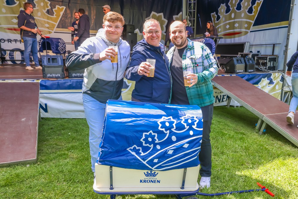 Drei Männer stehen lächelnd auf einer Wiese und halten Biergläser, neben ihnen ein gekühlter Bierwagen.