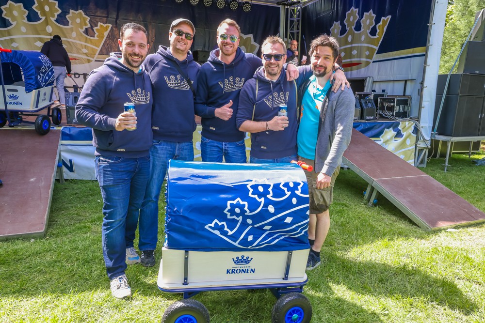 Fünf Männer lächeln neben einem Bollerwagen bei einem Outdoor-Event.