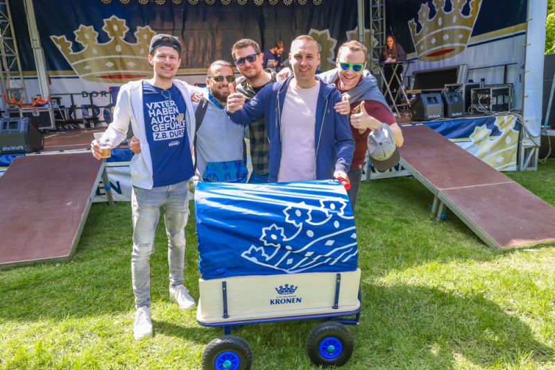Fünf Männer stehen lächelnd mit einem blauen Bollerwagen bei einem Festival.