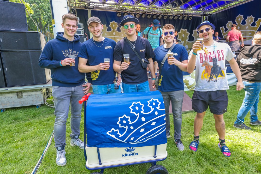 Fünf junge Männer stehen lächelnd vor einer Bühne und halten Getränke, neben ihnen ein kleiner blauer Wagen.