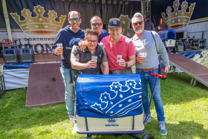 Fünf lächelnde Männer halten Gläser und stehen hinter einem Wagen auf einem Festival.