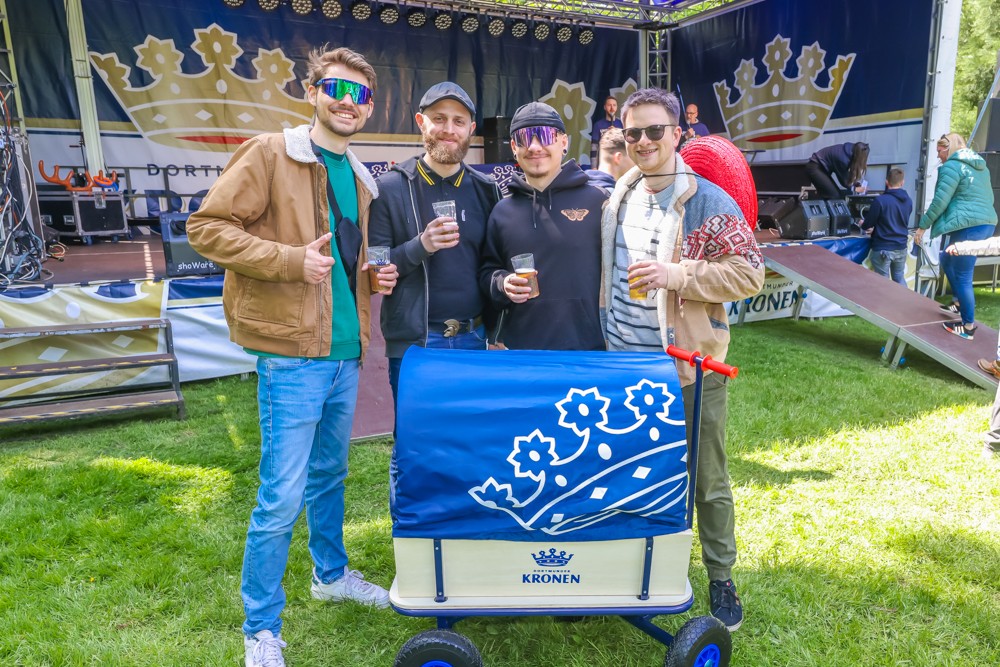 Vier Männer stehen mit Getränken neben einem Wagen auf einem Festival.