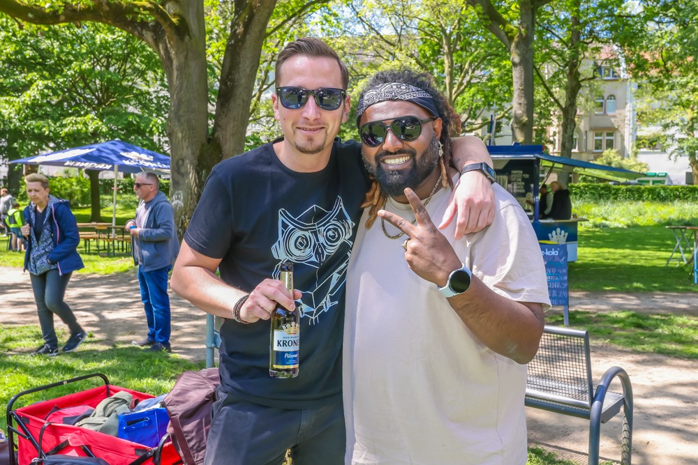 Zwei Männer stehen lächelnd in einem sonnigen Park und einer zeigt das Victory-Zeichen.
