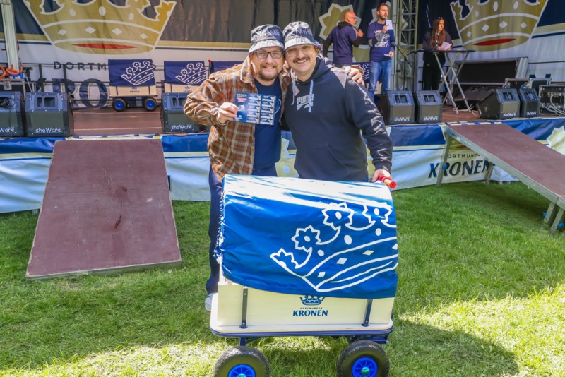 Zwei Männer stehen lächelnd vor einer Bühne, einer hält eine Kiste auf einem blauen Wagen.