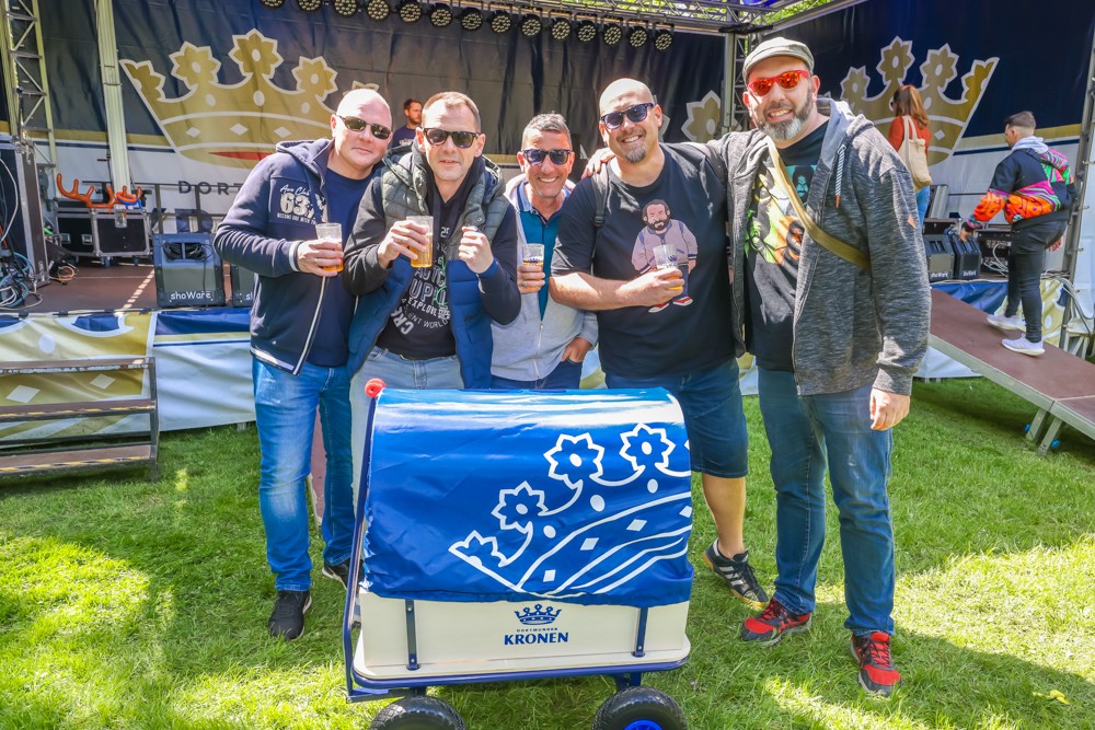 Fünf Männer stehen lächelnd auf einer Wiese und halten Biergläser. Sie stehen neben einem Getränkewagen.