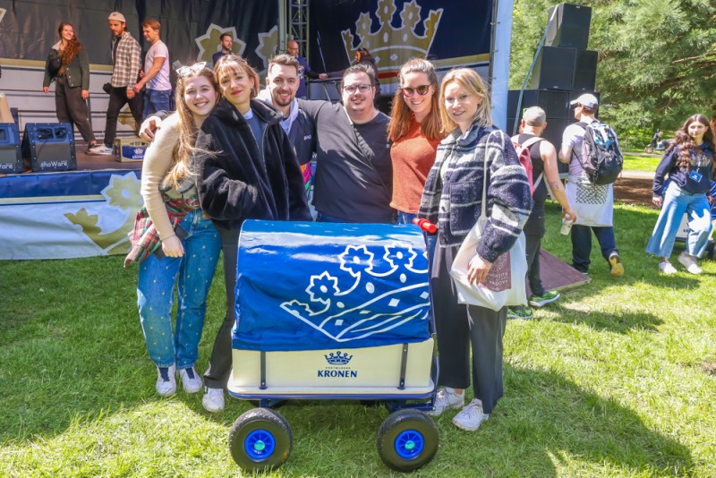 Sechs Personen stehen neben einem Bollerwagen in einem Park, Musikband spielt im Hintergrund.