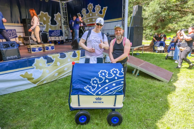 Zwei Männer stehen lächelnd vor einer Konzertbühne neben einem blauen Bollerwagen.