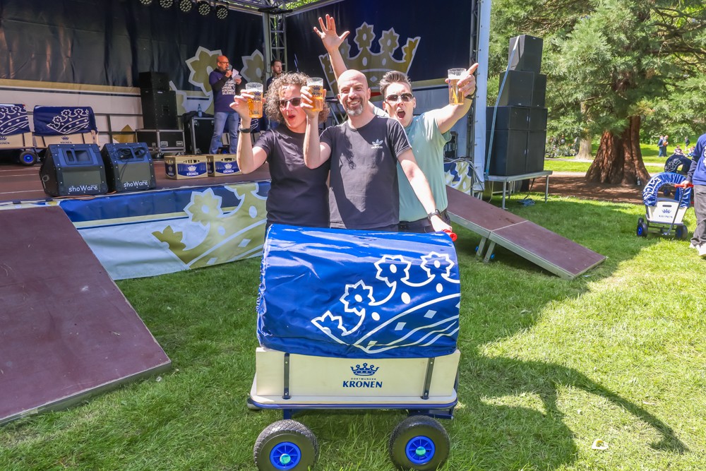 Drei Personen feiern mit Biergläsern in der Hand neben einer Bühne im Park.
