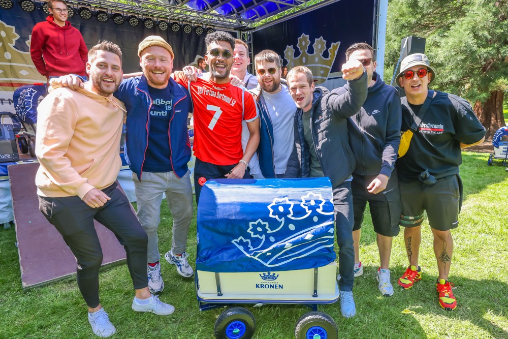 Acht Männer lächeln glücklich im Freien neben einem blauen Wagen.