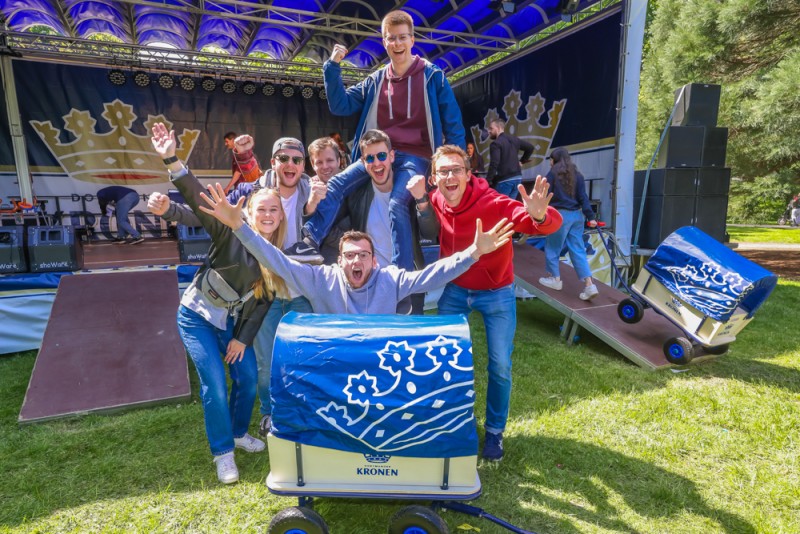 Sieben junge Erwachsene freuen sich auf einem Festival vor einer Bühne. Zwei halten einen blau dekorierten Wagen.