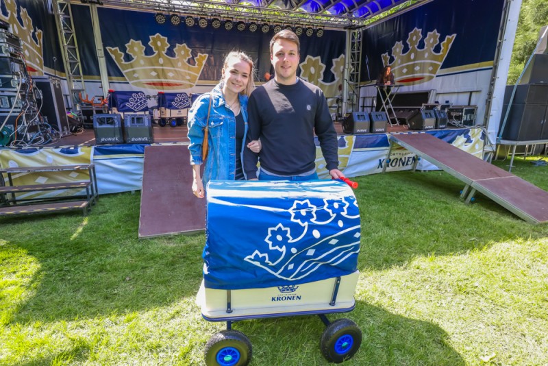 Zwei lächelnde Personen stehen mit einem blauen Bollerwagen vor einer Bühne.