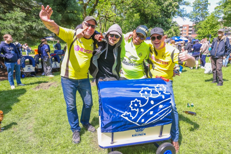 Vier fröhliche Erwachsene in farbigen Sporttrikots posieren mit einem blauen Bollerwagen im Park.