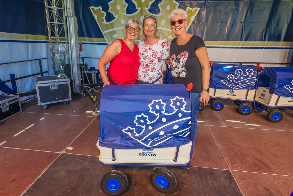Drei lächelnde Frauen stehen neben blauen Handwagen auf einer Bühne.