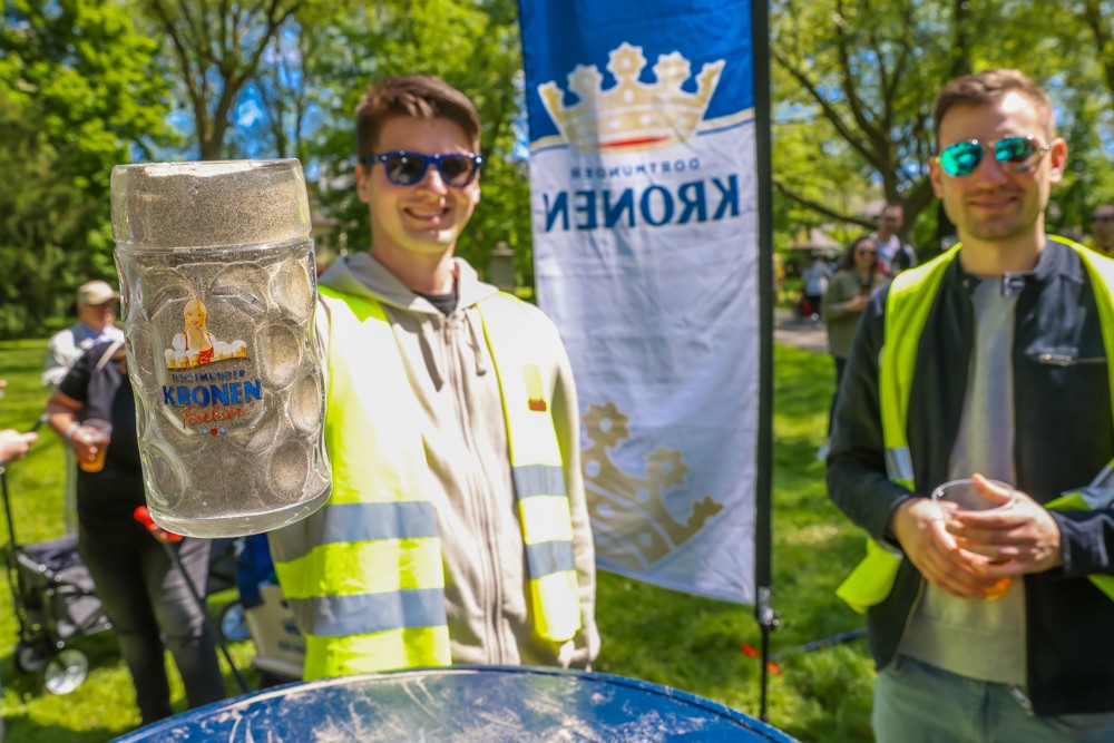 Zwei Männer präsentieren im Freien ein großes gefülltes Bierglas. Sie stehen neben einer blauen Fahne.