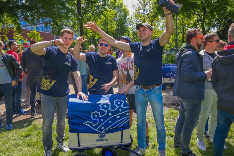 Vier Männer stehen lächelnd im Freien, feiern und schieben einen Bollerwagen.