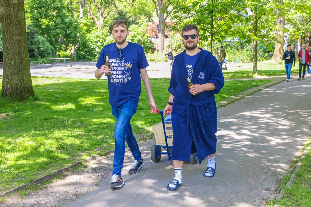 Zwei Männer gehen in einem Park, einer trägt einen blauen Bademantel, der andere ein blaues T-Shirt.