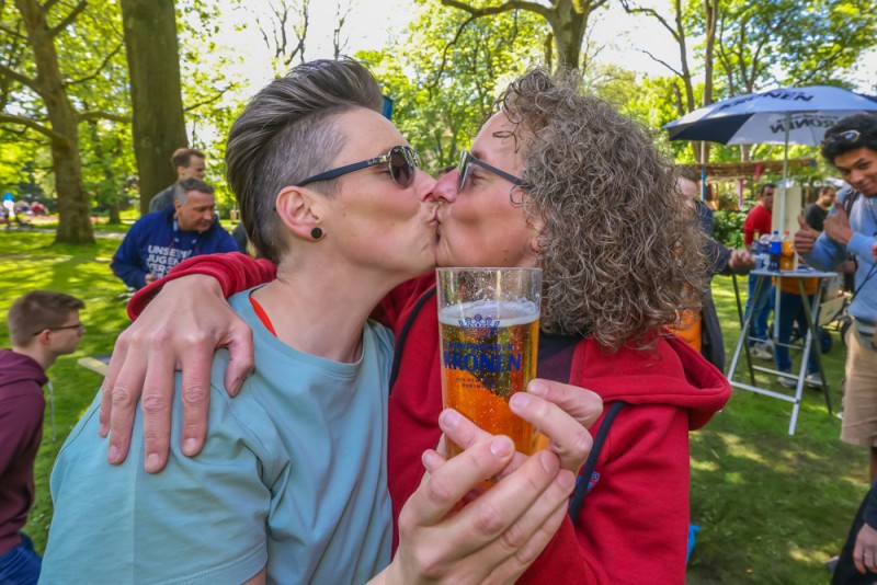 Zwei Frauen umarmen sich und küssen sich in einem belebten Park. Eine hält dabei ein Bierglas.
