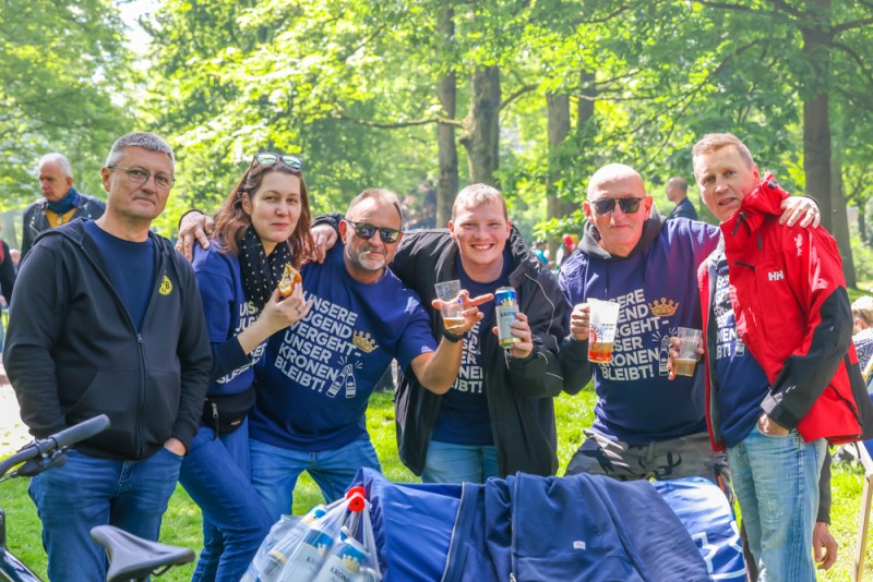 Sechs Personen posieren lächelnd im Park, einige halten Getränke. Sie tragen blaue und dunkle Kleidung.