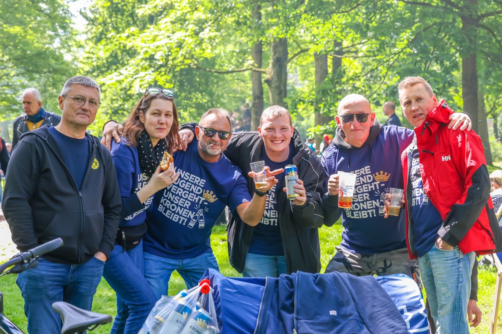 Sechs Personen posieren lächelnd im Park, einige halten Getränke. Sie tragen blaue und dunkle Kleidung.
