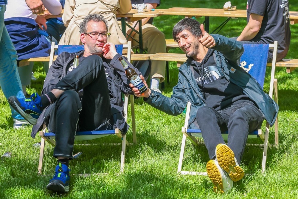 Zwei Männer sitzen entspannt auf Klappstühlen im Park und unterhalten sich bei sonnigem Wetter.