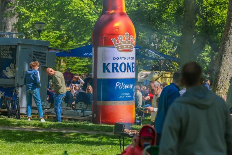 Eine große aufblasbare Flasche steht in einem belebten Park neben Menschen und Essensständen.