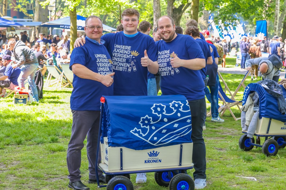 Drei Männer in blauen T-Shirts stehen neben einem Bollerwagen in einem Park.
