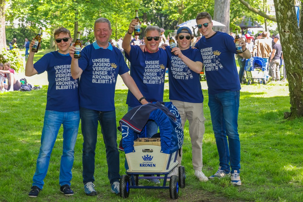 Fünf lächelnde Männer in blauen Shirts halten Getränke und stehen im Park.